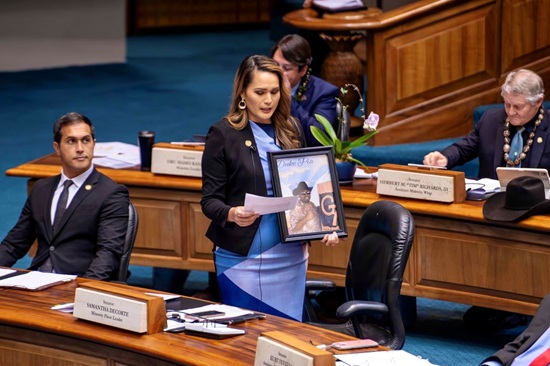 Senator Samantha DeCorte standing at her desk on the Hawaii State Senate floor, holding a piece of paper and a framed portrait, with a nameplate on her desk identifying her as a senator and minority floor leader